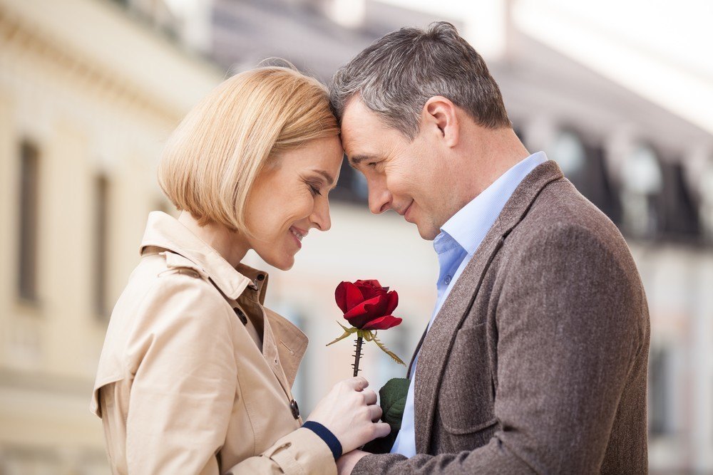 Portrait of two people holding rose and smiling. adult man giving red flower to blond woman outside